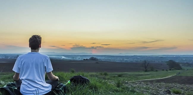 Jeune homme de dos assis dans l'herbe admirant un paysage lointain et un coucher de soleil