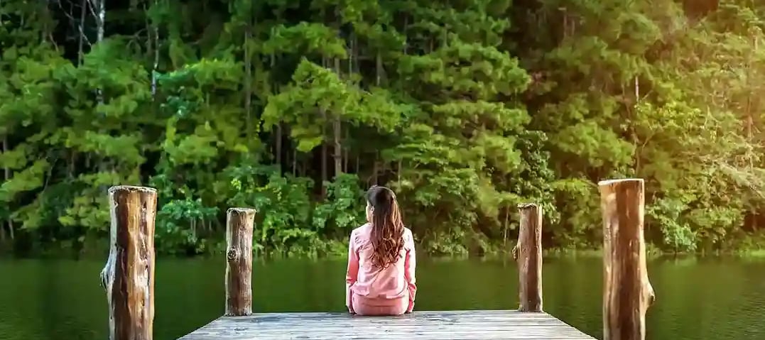 Femme de dos cheveux longs habits roses assise sur un ponton au bord d'une rivière bordée d'arbres