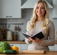 A smiling woman in a kitchen holding a notebook, surrounded by fresh vegetables.