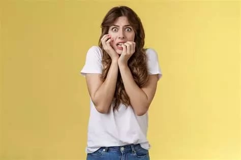 Femme blonde avec visage effrayé bras droit sur joue et doigts main gauche dans la bouche