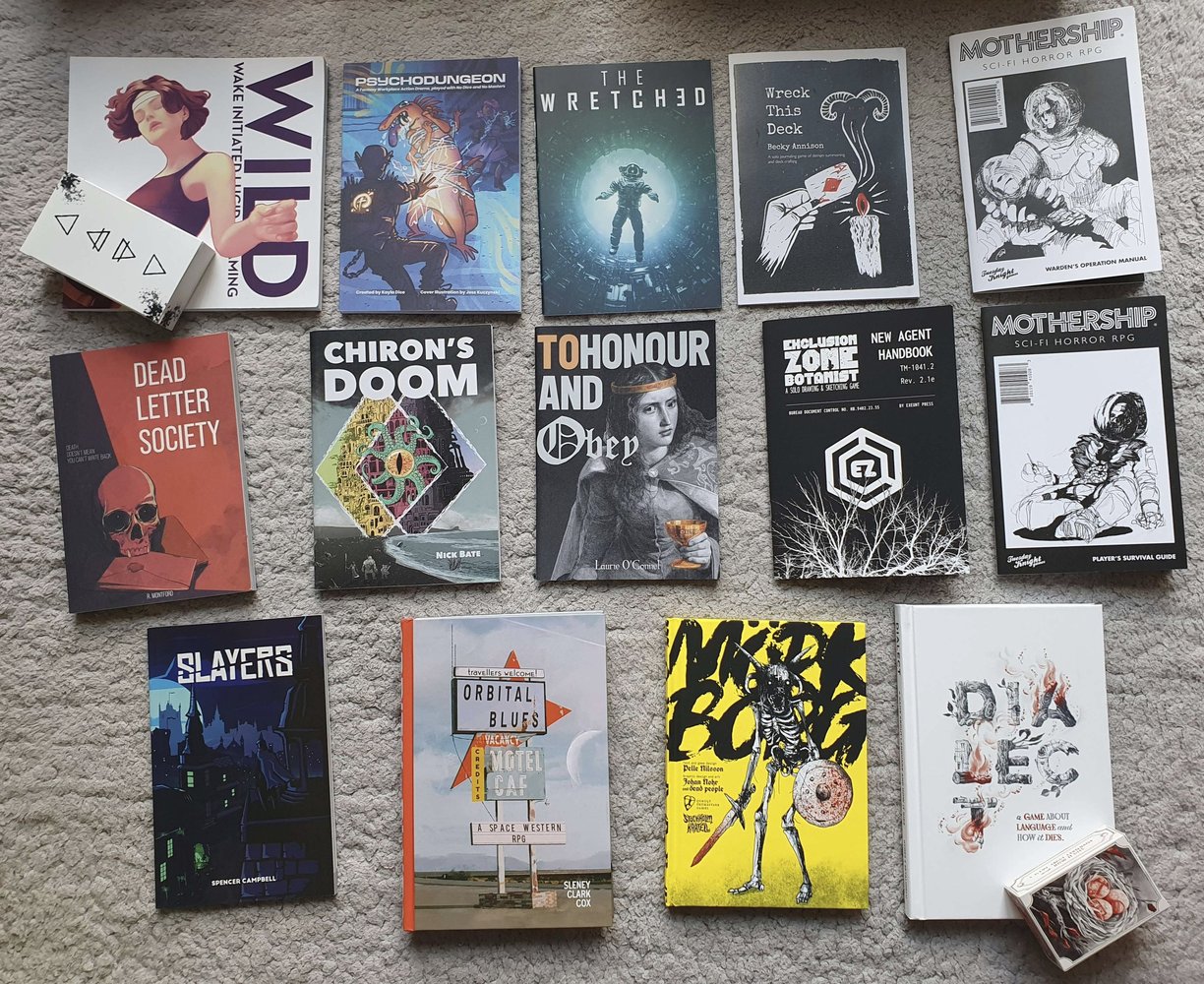 A selection of TTRPGs, mostly zines, laid out on a carpet in rows.