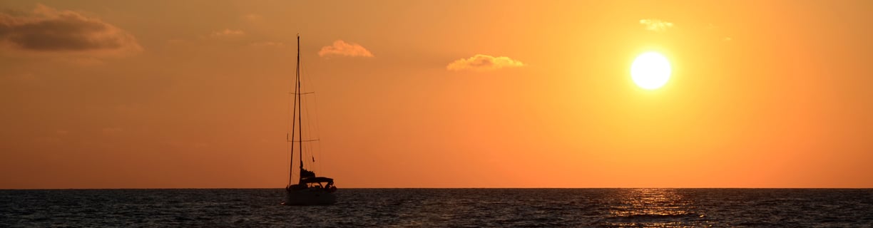 Horizonte durante el atardecer, con el cielo naranja, y el sol y un velero de fondo en el mar