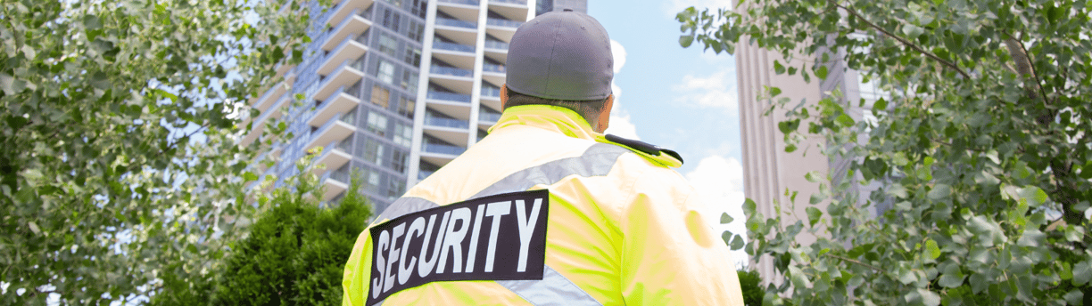Ein Sicherheitsmitarbeiter mit einer "SECURITY"- Jacke mit dem Blick auf ein Hochhaus.
