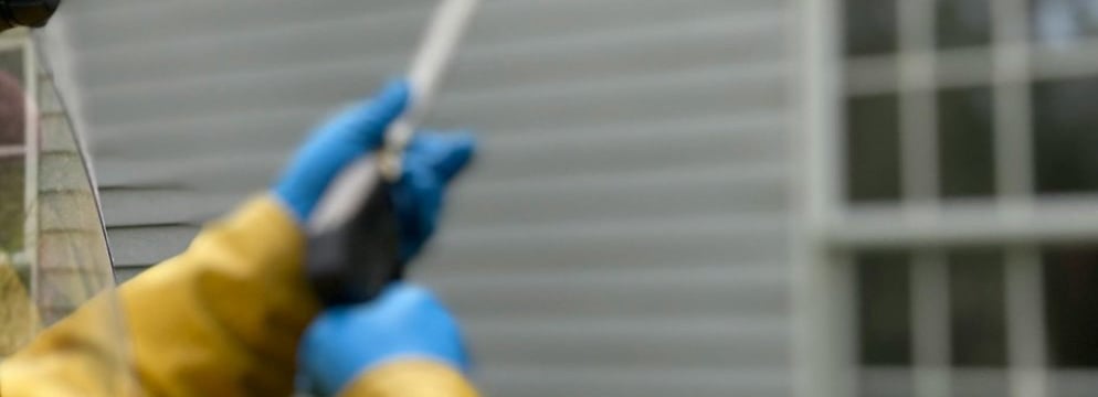 hands in blue plastic gloves hold a pressure washing lance pointing at a house wall in background