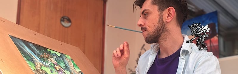 A handsome young male artist with dark hair and blue shirt sits at his easel of fantasy art