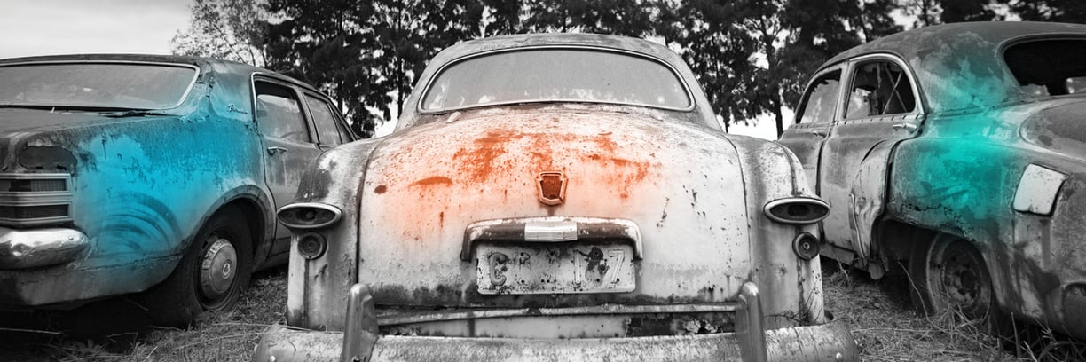 Three abandoned vintage cars in a field – a turquoise sedan flanked by rusty companions beneath shadowy trees.