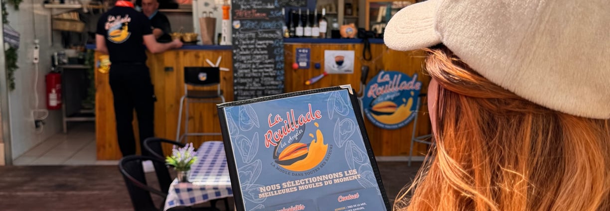 Une jeune femme lit le menu de La Rouillade lors d'une journée ensoleillée.