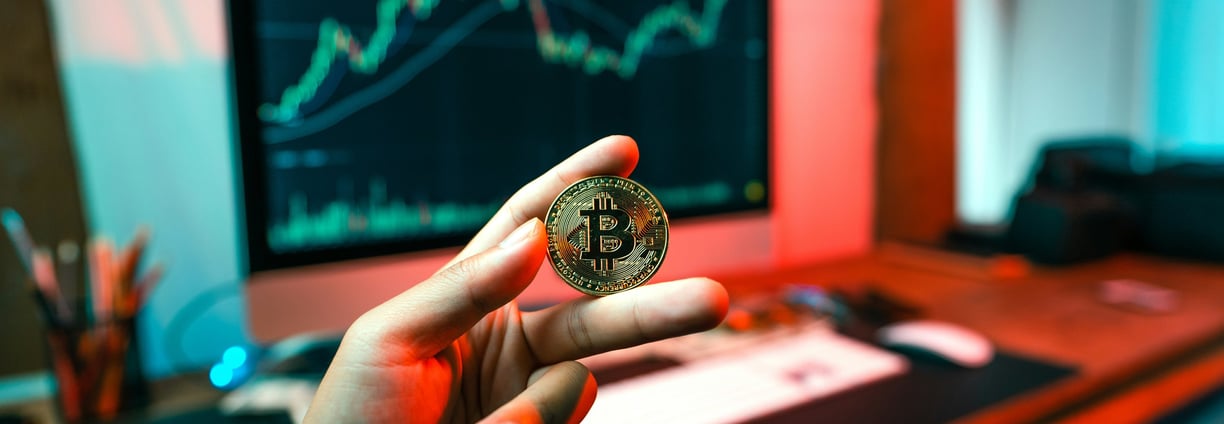 A person holds a bitcoin in front of a computer screen displaying cryptocurrency data and charts.