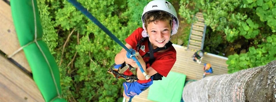 A person is zip-lining across a lush, green landscape, surrounded by dense foliage and rocky outcrop