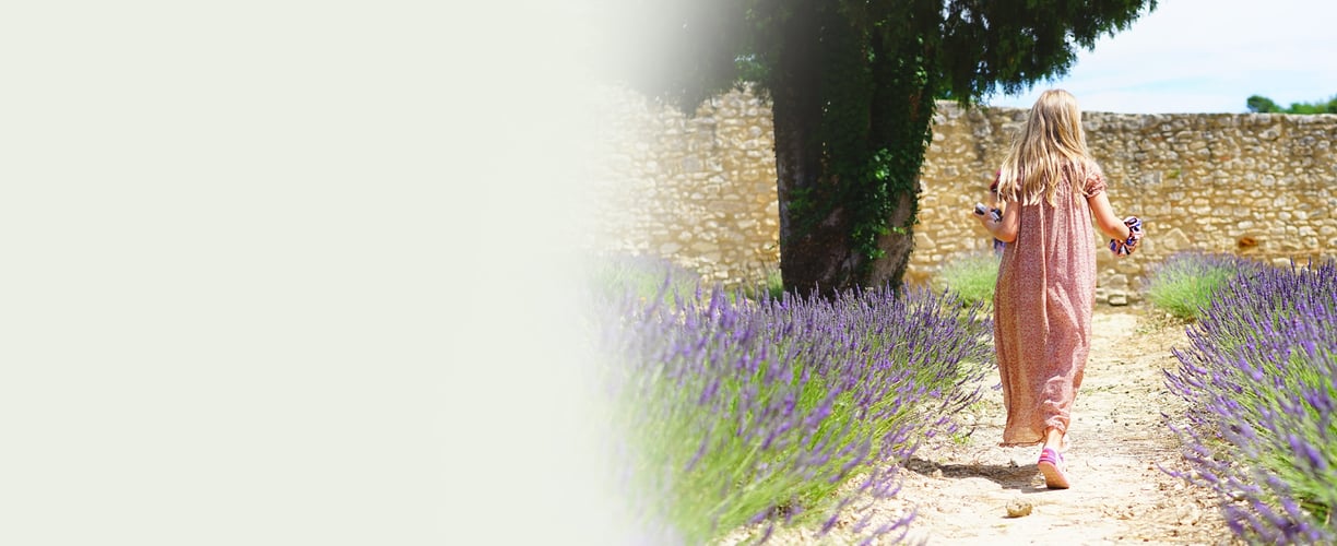 A young girl is wearing a long floral dress, walks away down a path through a lavender field
