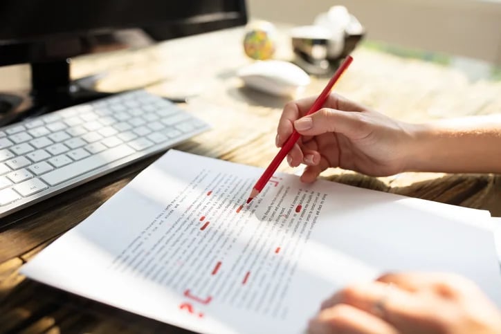 a person proofreading and correcting a document with a red pencil, computer in the background