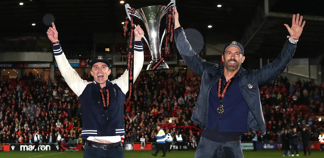 "Ryan Reynolds and Rob McElhenney celebrating on the football pitch, holding the Vanarama National L