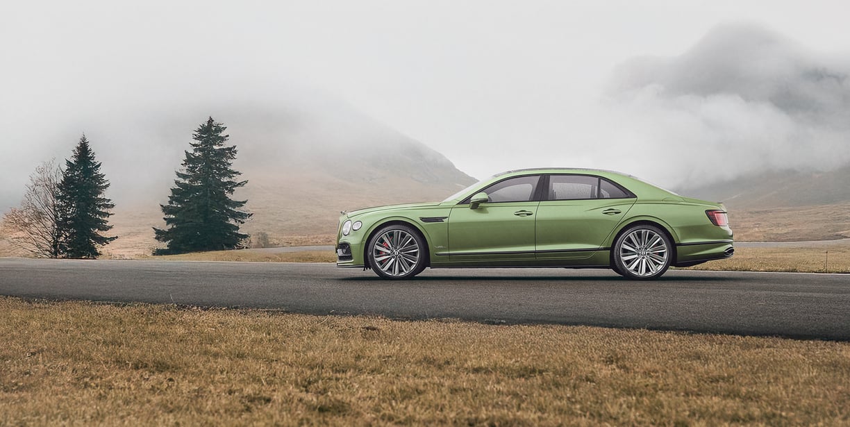 a green bentley driving down a road with a mountain in the background