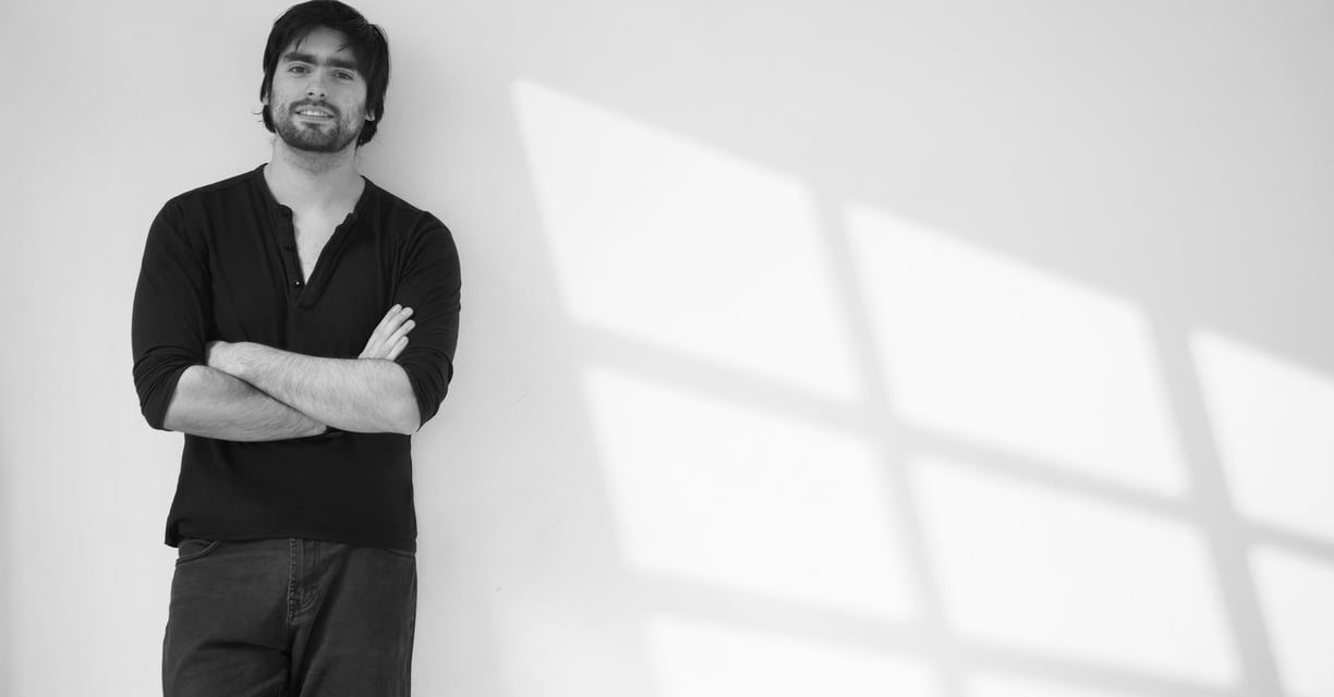 Hombre de cabello oscuro y barba, vestido de negro, apoyado contra una pared blanca con sombras geométricas de una ventana pr