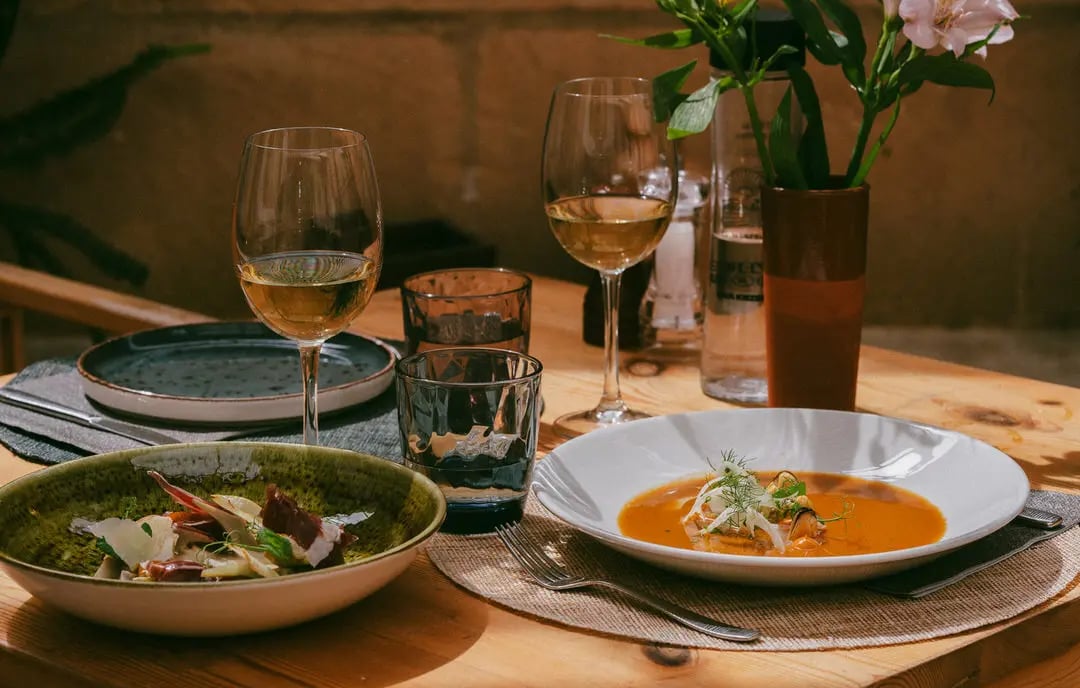 Mesa montada con comida encima, se ve deliciosa, alcachofas y sopa de pescado. Hay flores bonitas en el restaurante Es Mestra