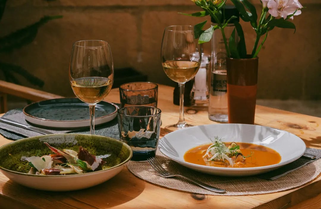 Mesa montada con comida encima, se ve deliciosa, alcachofas y sopa de pescado. Hay flores bonitas en el restaurante Es Mestra