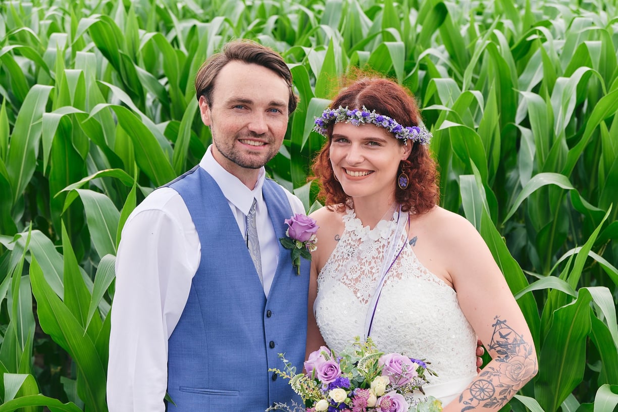 a bride holding a bouquet and a groom standing next to each other in a corn field