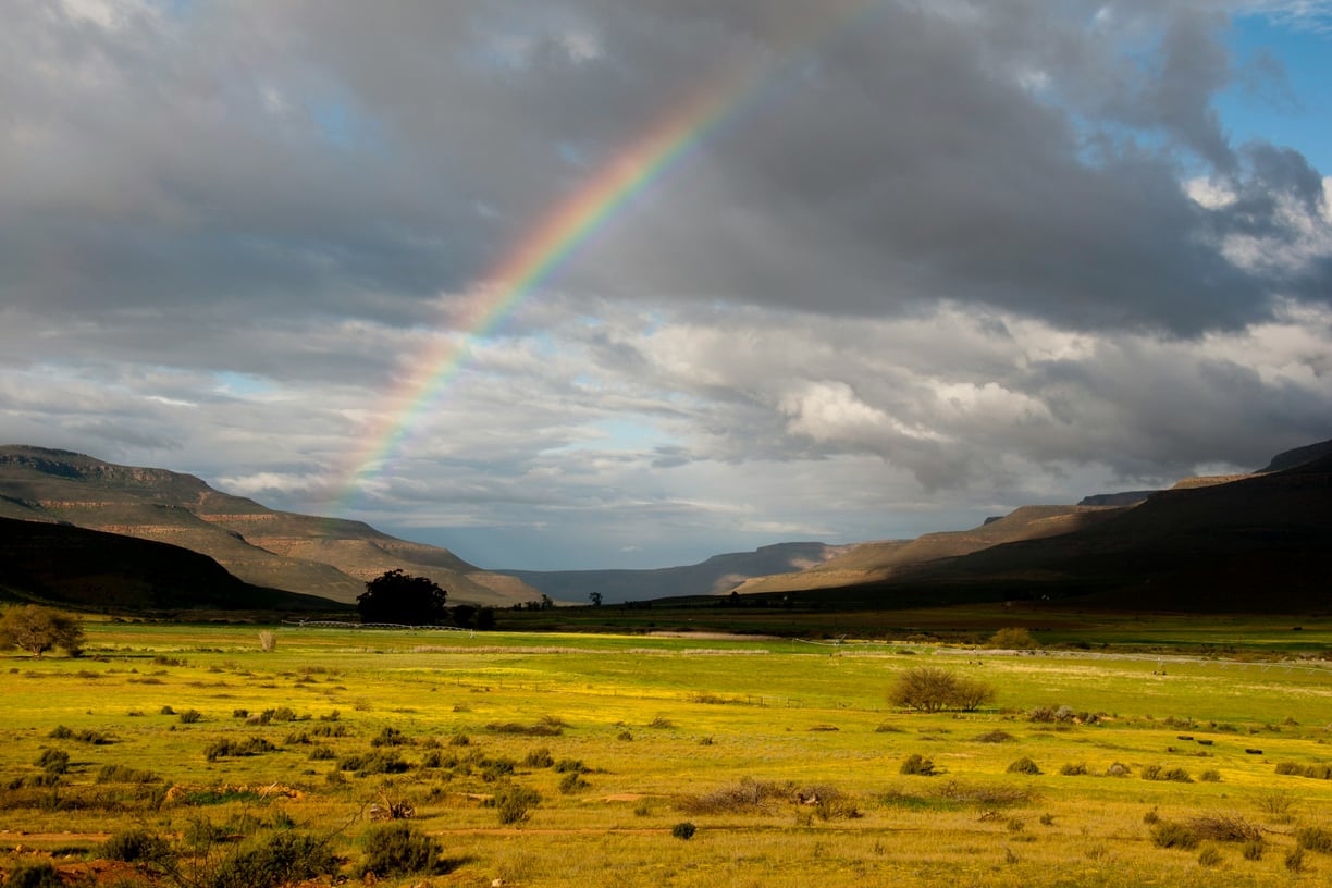 Rainbow across a cloudy sky with some fields in the foreground and a circular mountain range as backdrop