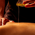 A hand pours oil from onto the back of a lying woman, another hand rests on the woman's shoulder