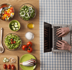 Person typing on laptop at other end of table that displays many dishes/ingredients