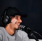 aledima sorride mentre parla al microfono durante un podcast, con cuffie e cappello nero