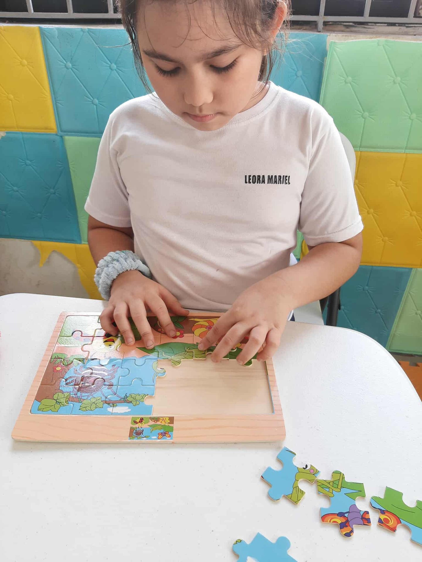 A young girl in a white t-shirt focuses on assembling a colorful wooden animal puzzle at a white table.