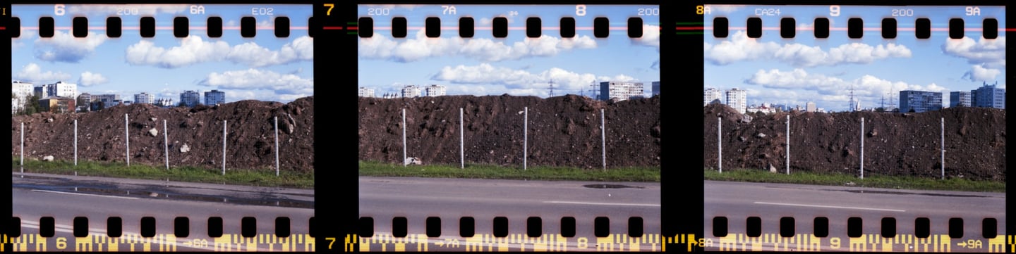 Composed photo of a street with a large, dark, rocky mound, cityscape in the background and clouds on the blue sky