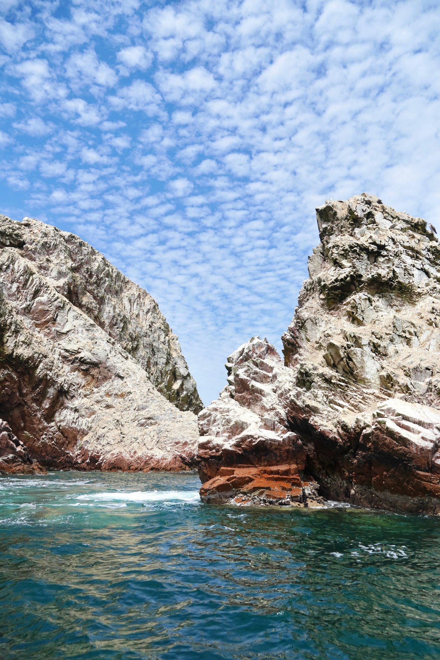 Rugged coastal rock formations in the Ballestas Islands, Peru, under a bright blue cirrocumulus sky.