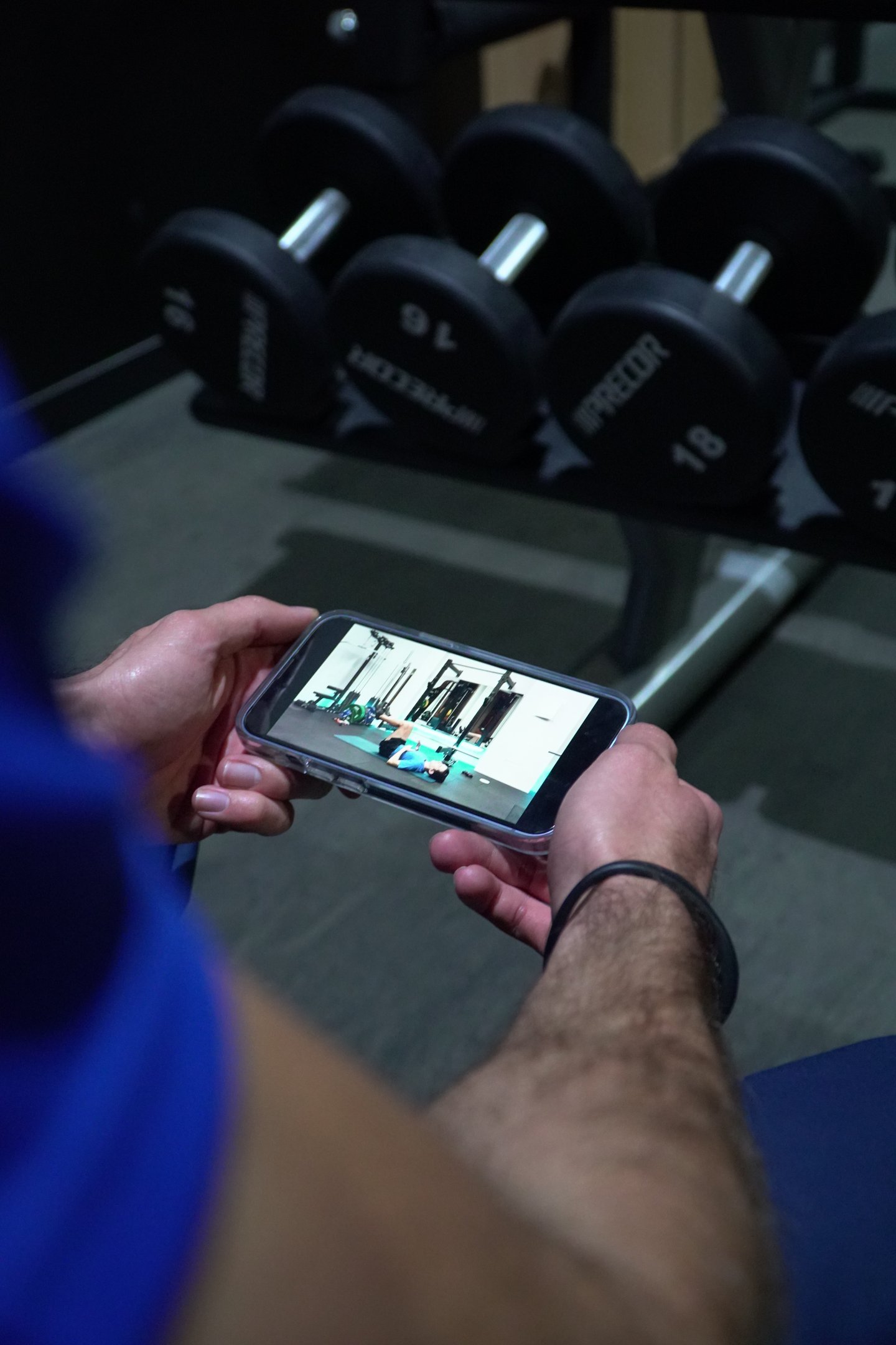 Person in a gym holding a smartphone watching an online fitness workout video near dumbbells.