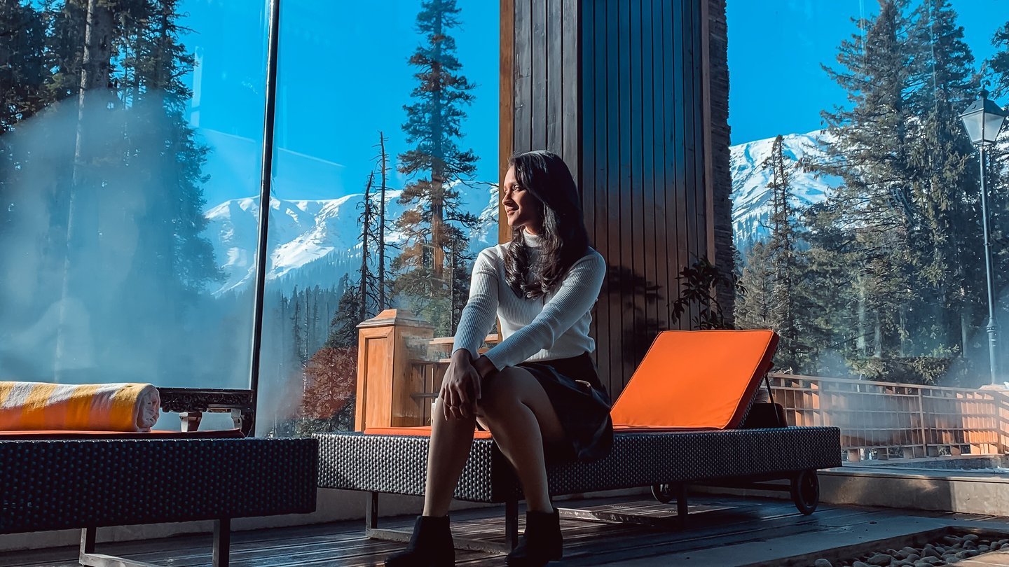 A woman relaxes on a lounge chair at a mountain resort with a scenic view of snow-capped peaks and pine trees.