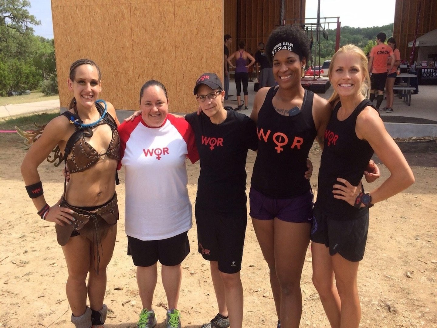 Five diverse female athletes smiling together at an outdoor OCR mud run event wearing WOR team shirts.