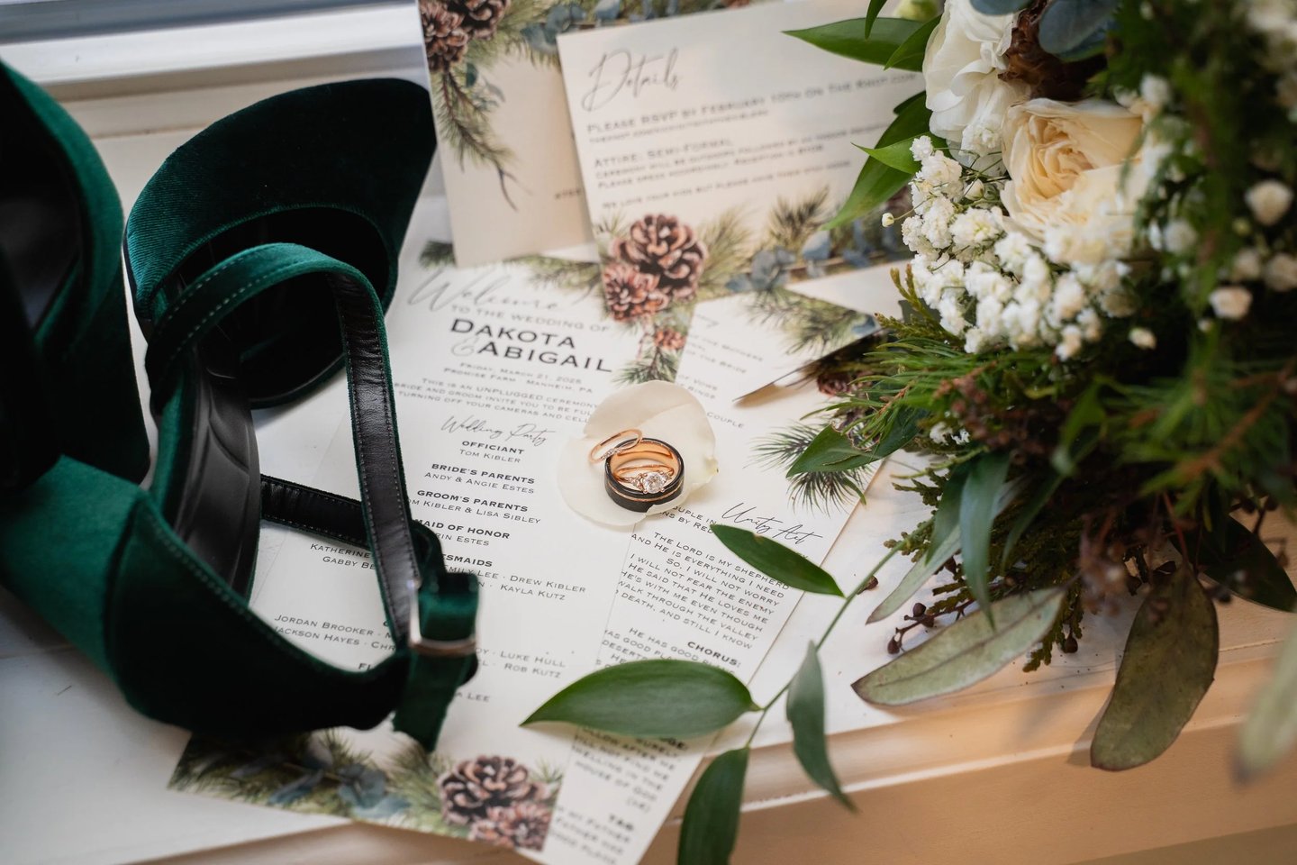 Emerald green velvet bridal heels, wedding rings, and stationery with a winter pine bouquet.