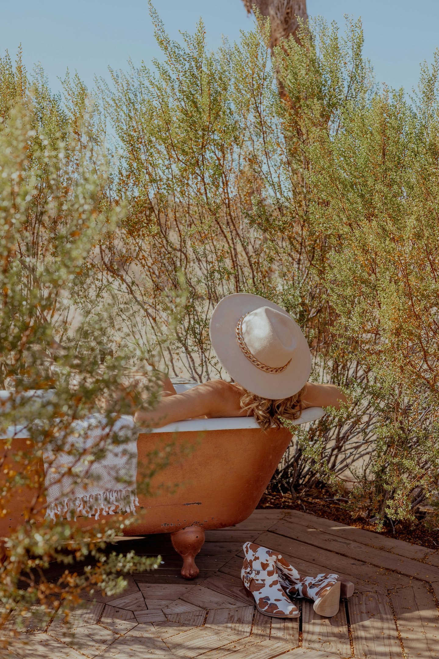 A woman in a wide-brim hat relaxes in an outdoor copper bathtub surrounded by desert greenery.