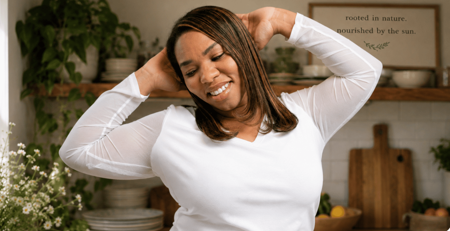 Smiling woman in a white long-sleeve shirt stretching in a bright kitchen with plants.