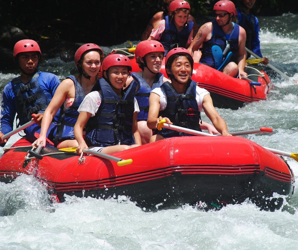 A group of tourists wearing red helmets and life jackets white water rafting on a red inflatable boat.