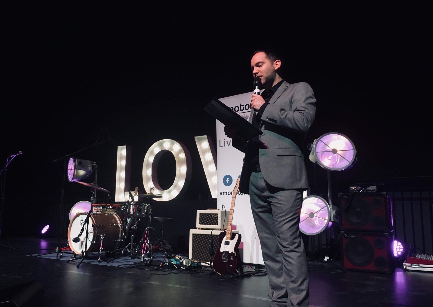 Alex hosting an awards function in a grey suit holding a clipboard
