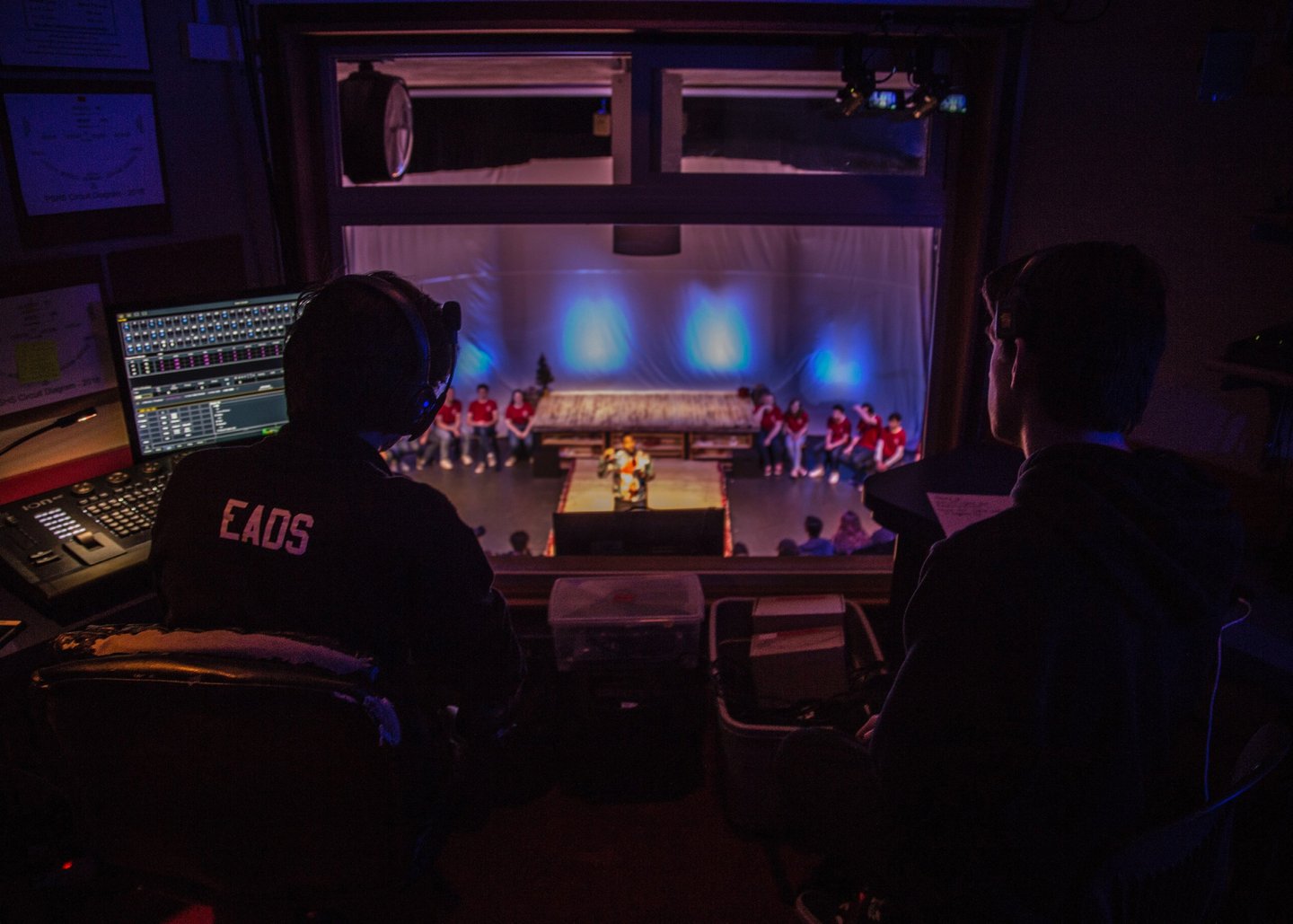 Two figures facing away from the camera looking through a window to the stage