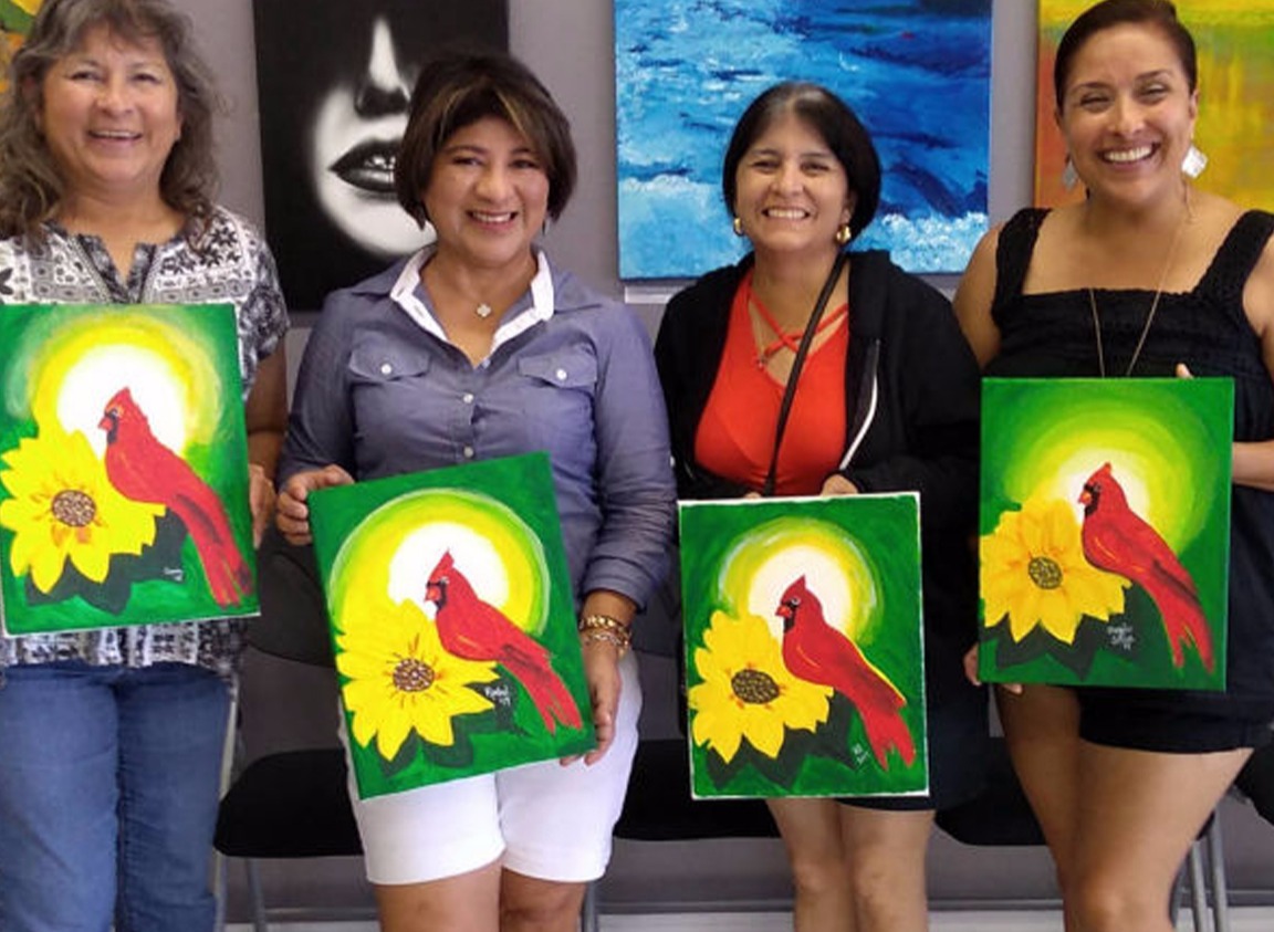 a group of smiling women holding up their paintings of a cardinal and a sunflower