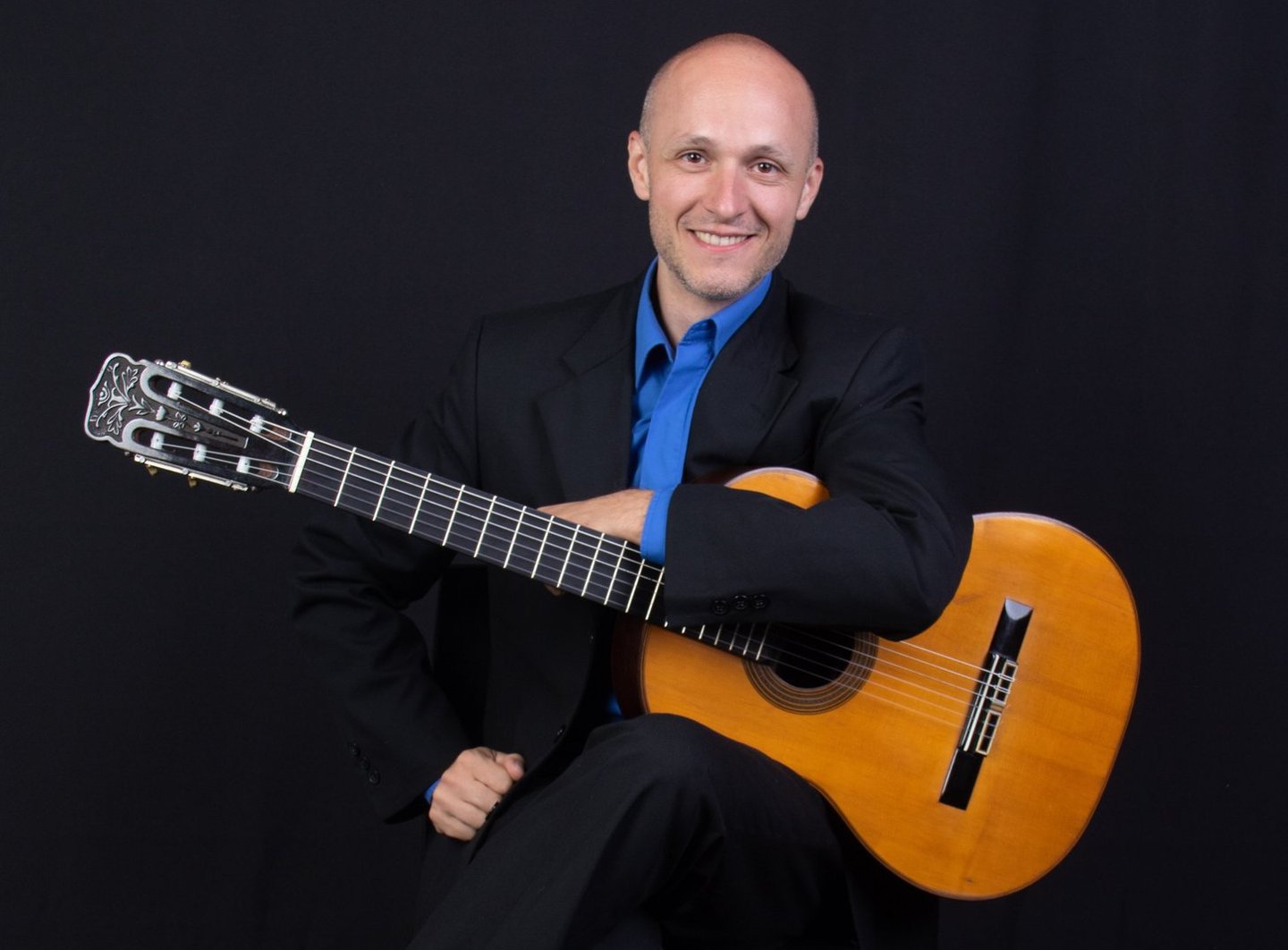 Antonio Gomez con ambo negro y camisa azul francia sentado con su guitarra en las manos