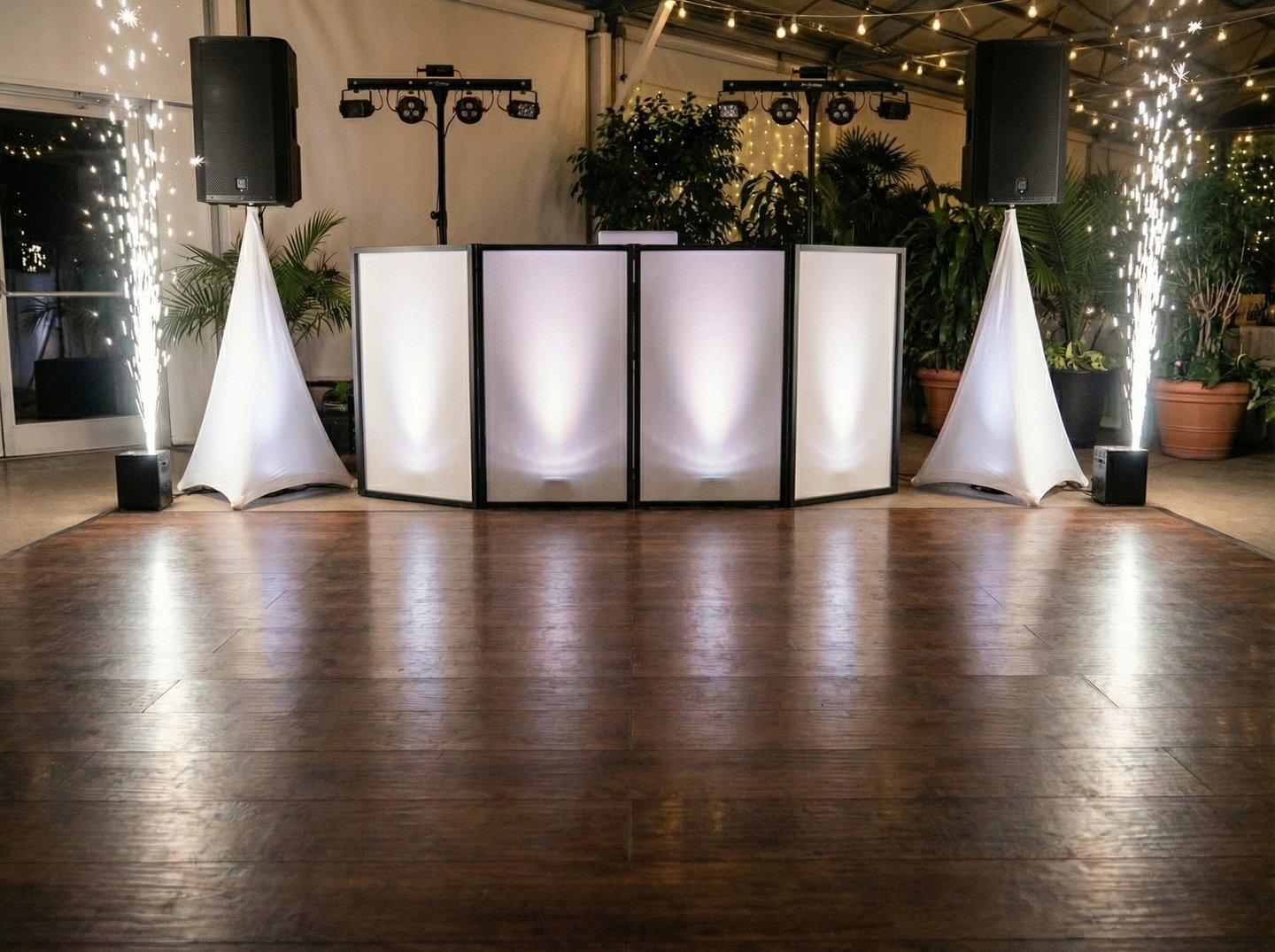 Professional wedding DJ booth setup with white facade, speakers, and cold sparklers on a wood dance floor.