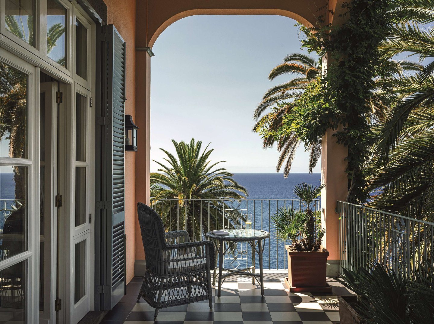 Luxury hotel balcony with ocean views, palm trees, and wicker furniture on a sunny day.