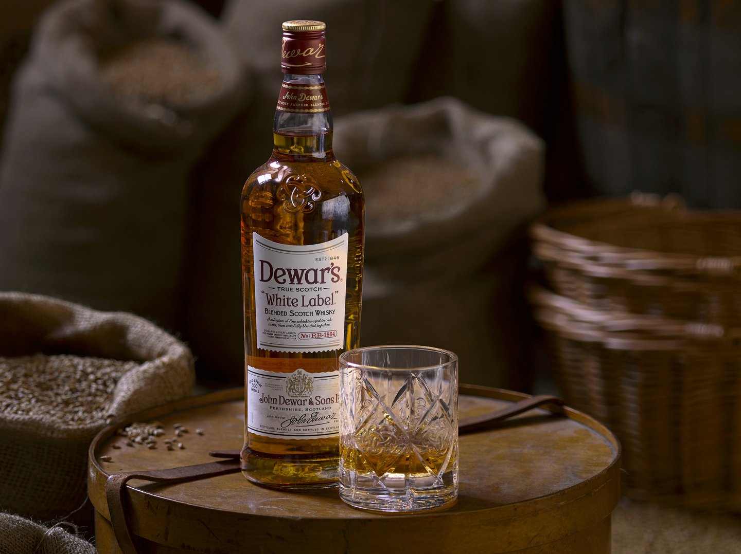 Dewars whisky bottle and glass on a round wooden box next to open sacks of grain 