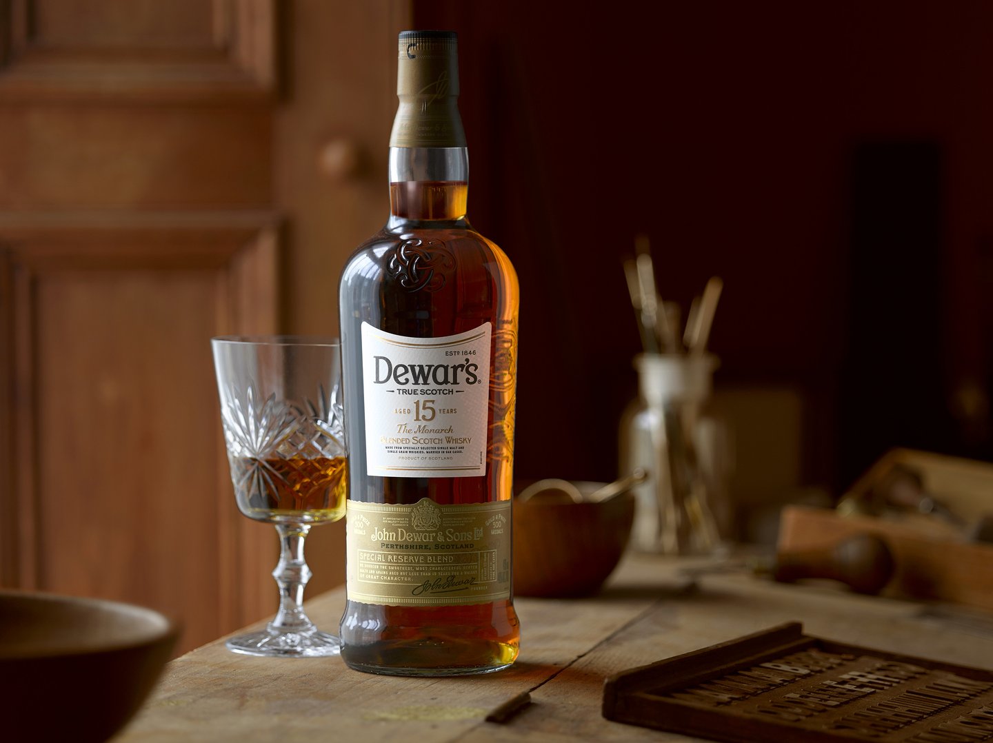 Dewars whisky bottle next to glass of whisky on a wooden table next to a window