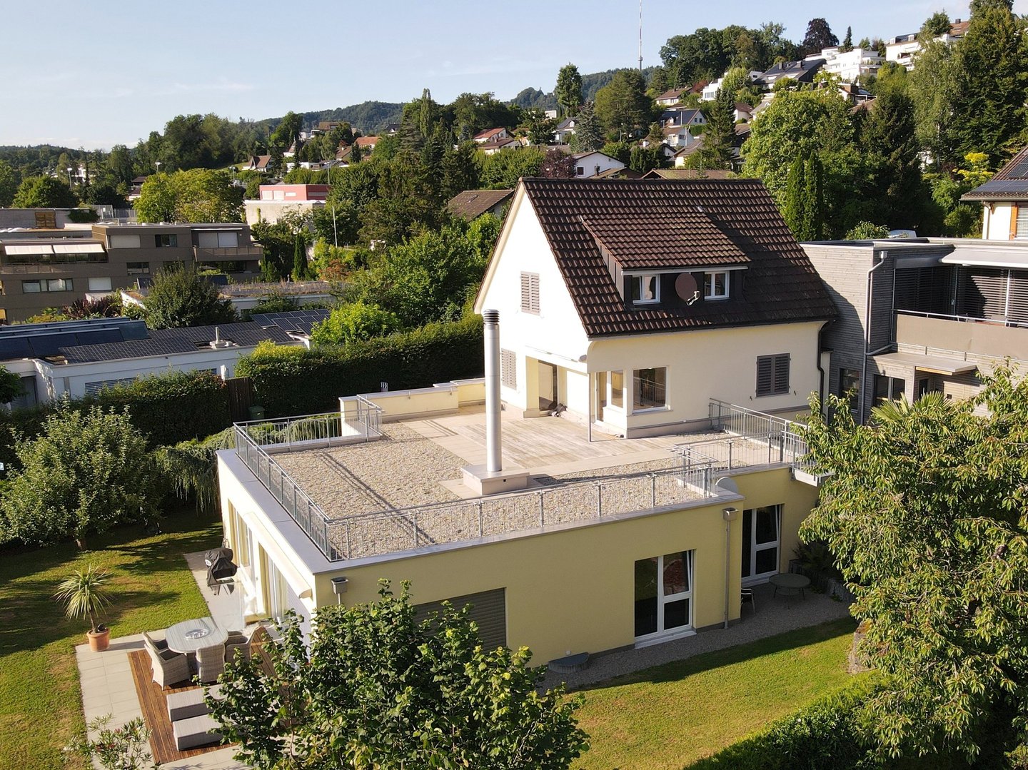Luftaufnahme eines Vorstadthauses mit großer flacher Dachterrasse und grüner Gartenlandschaft.