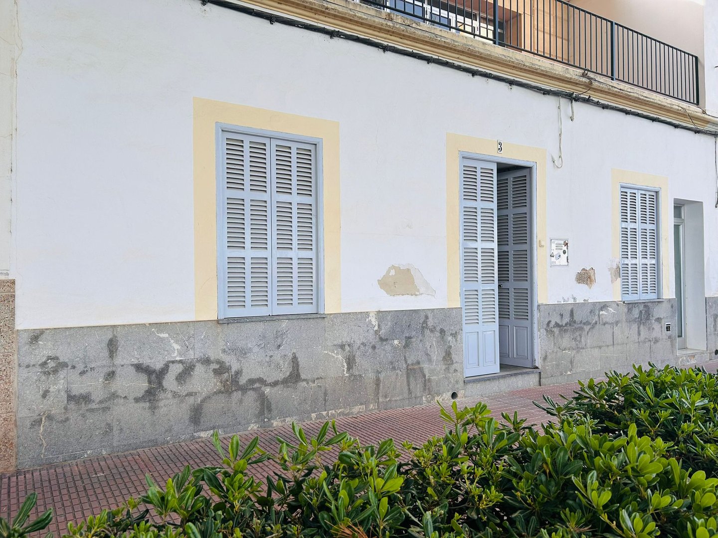 Traditional white Mediterranean house facade featuring light blue wooden shutters and a sidewalk with green bushes.