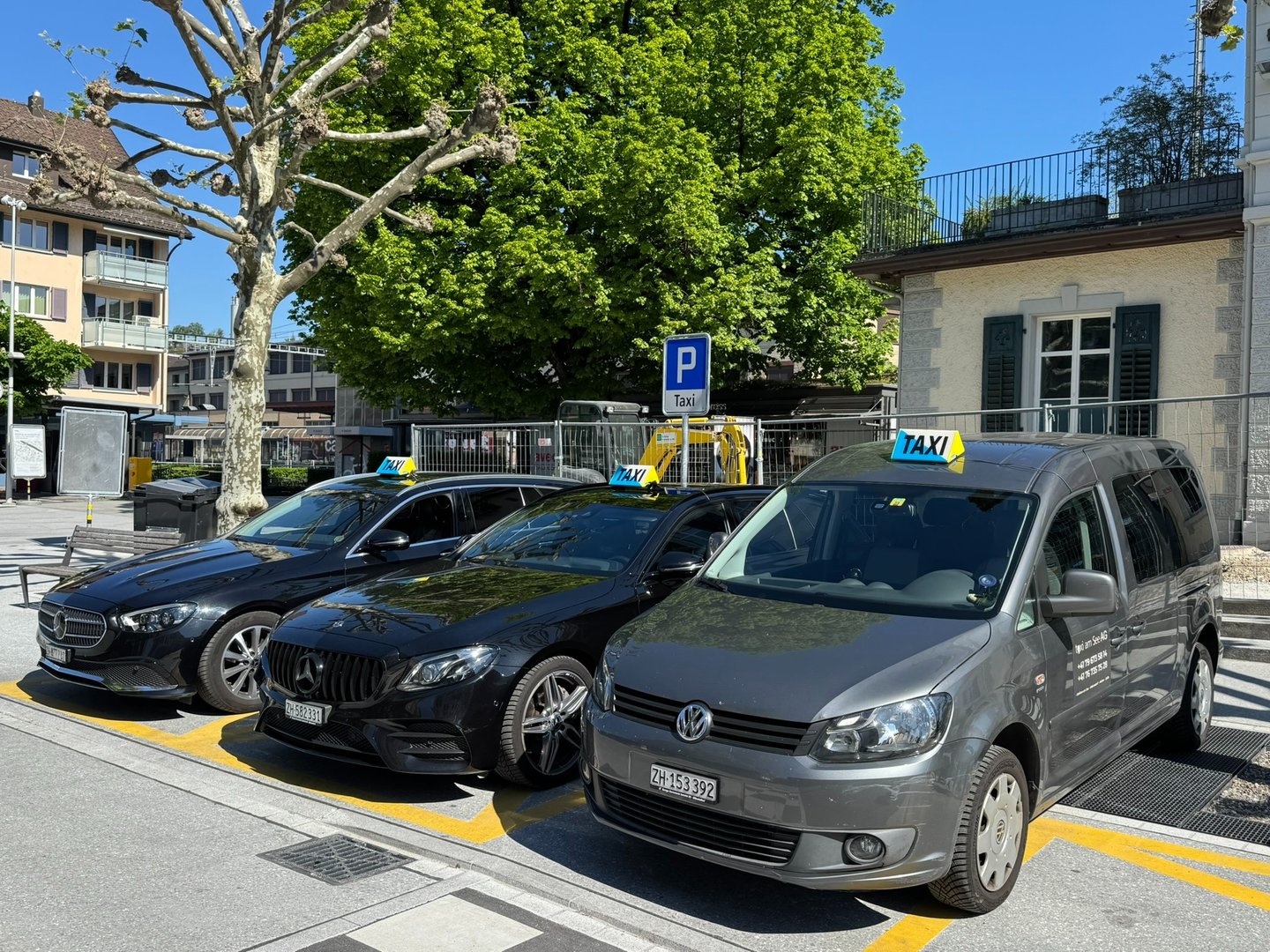 Three taxicars. One grey VW Caddy and two black Mercedes E-Classes parked on a swiss mainstation.