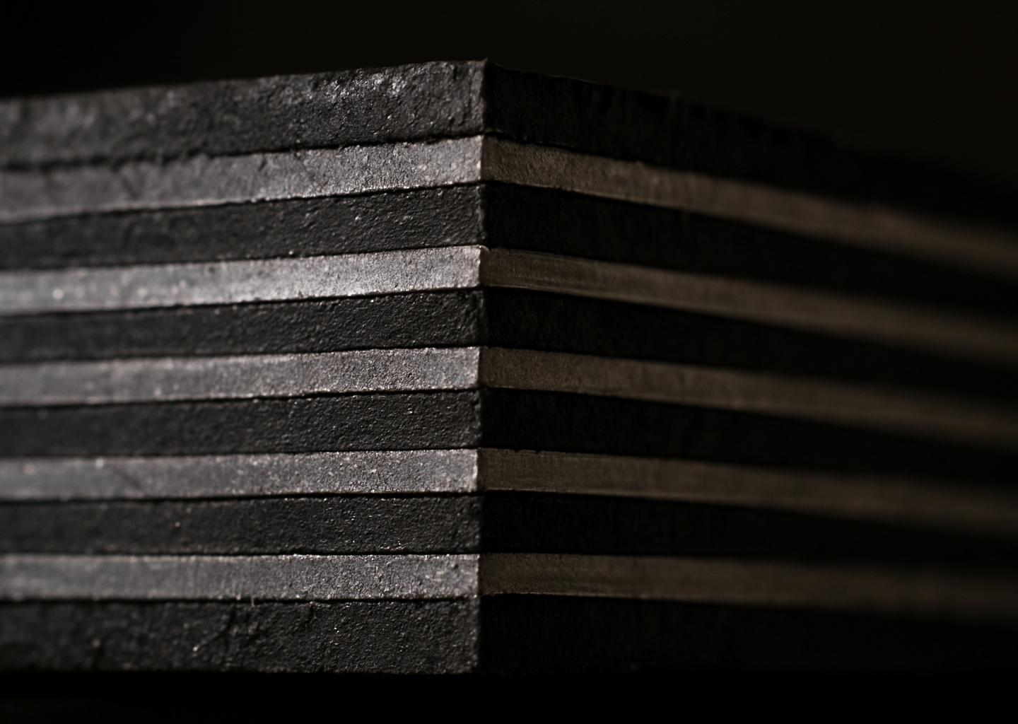 Extreme close-up cross-section of a laminated neoprene elastomeric bearing pad cut cleanly to reveal alternating black rubber layers and bright steel shim plates, harsh industrial workshop lighting from the side casting sharp shadows that emphasize the precision of each laminate layer, raw cut surface showing material density and bonding quality