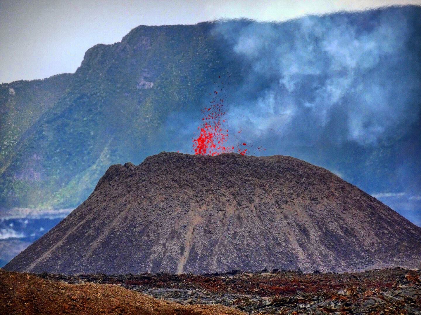 Red hot lava erupting from a volcanic cinder cone crater with smoke and mountain landscape.