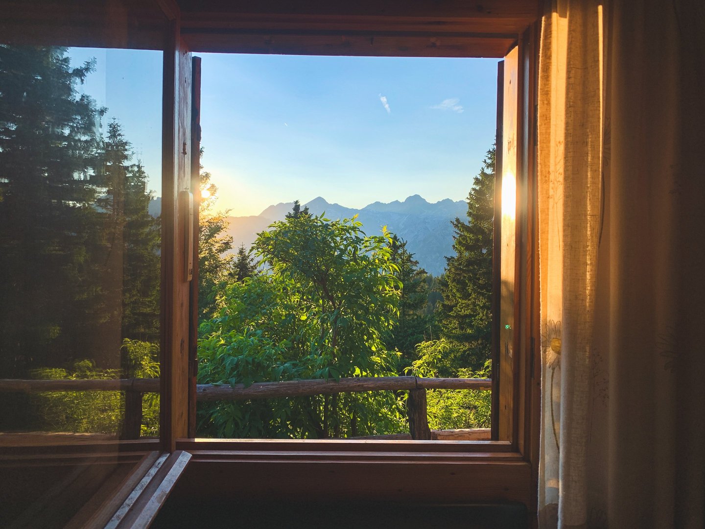 A scenic mountain sunset view through an open wooden cabin window surrounded by pine trees.