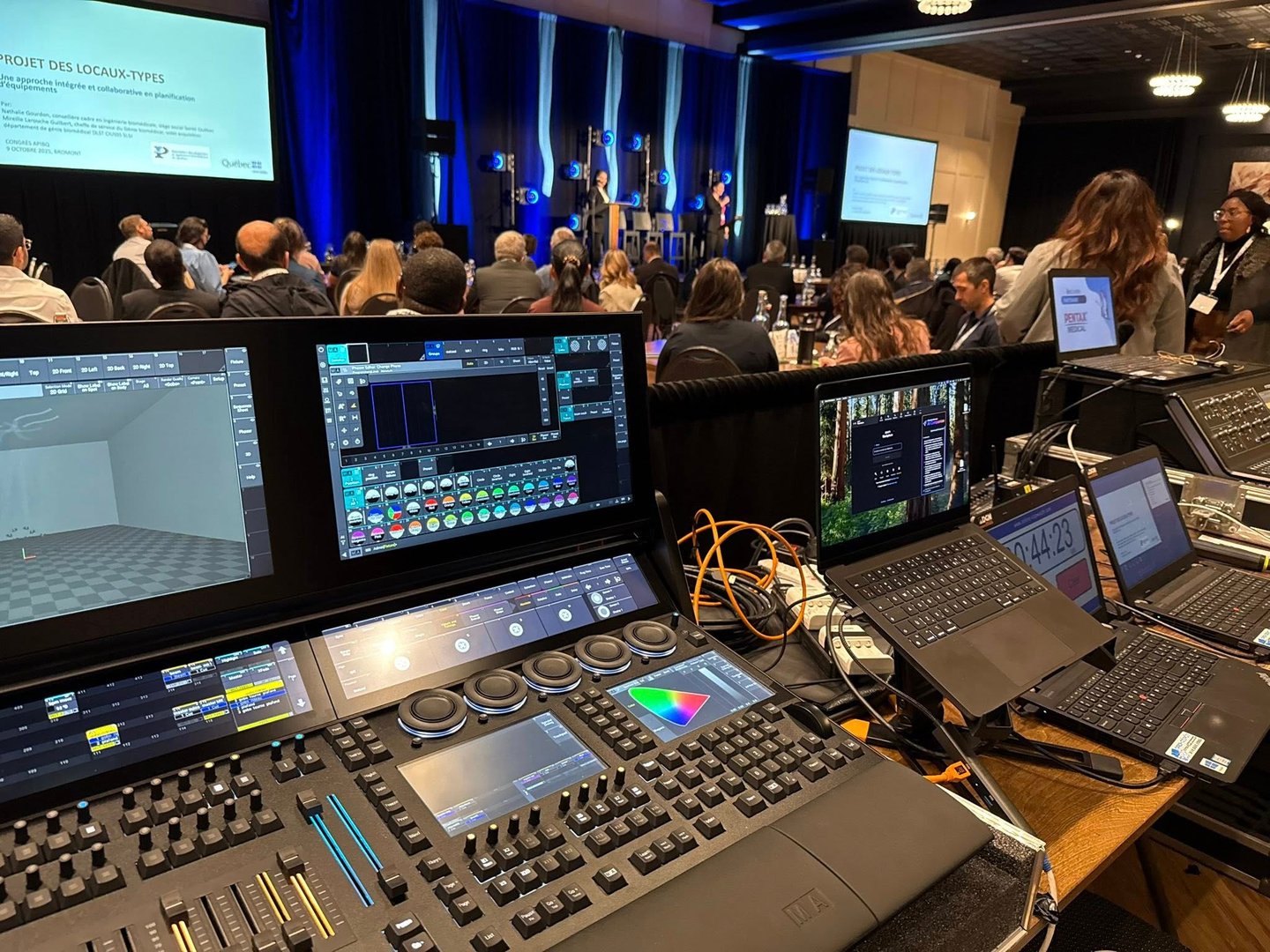 Professional lighting console and laptops at a live event production booth in a conference hall.